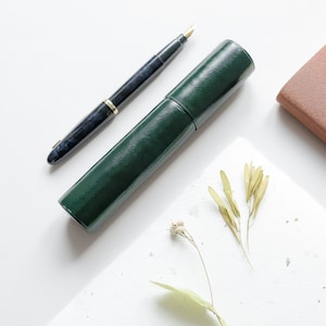 May include: A dark green leather pen case with a black and silver fountain pen. The pen case is lying on a white surface with a few small white flowers and green leaves.