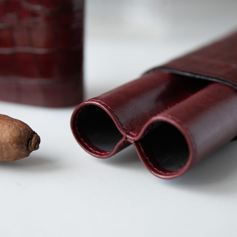 May include: A close-up of a burgundy leather cigar case with two openings. The case is next to a cigar and a matching burgundy leather container. The case has a textured surface and a black interior.