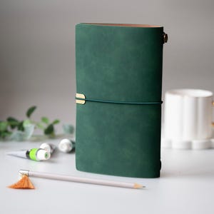 Puede incluir: Un diario de cuero verde oscuro con un cierre dorado y un cierre elástico a juego. Un lápiz con una borla naranja y tubos de pintura están en primer plano. Una taza blanca está en el fondo.