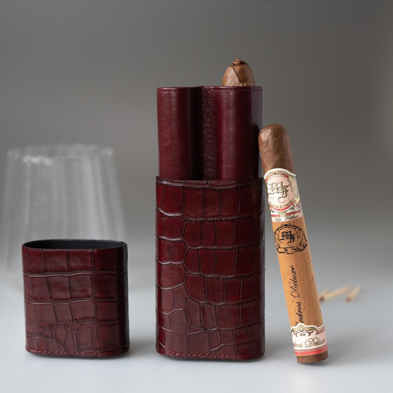May include: A burgundy leather cigar case with a crocodile skin texture, open to reveal a cigar. A lit cigar rests against the case. The case is on a white surface.