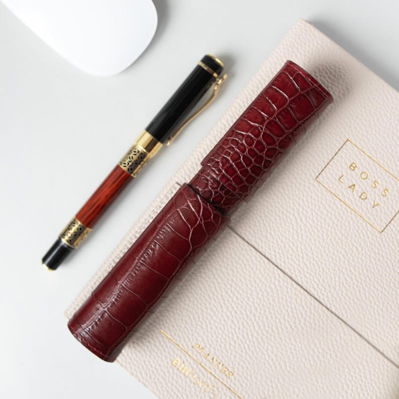 May include: A dark red, crocodile-textured cylindrical pen case resting on a beige planner with the text BOSS LADY embossed on the cover. Beside the case lies a black and wood-toned fountain pen with gold-colored accents and a clip.