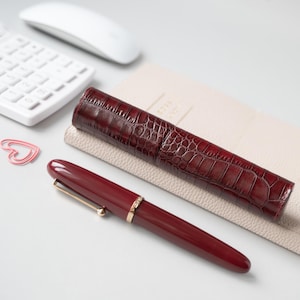 May include: A deep burgundy fountain pen with a gold-toned clip and band, accompanied by a matching cylindrical pen case featuring a dark red crocodile-embossed texture. The set is arranged alongside a white calculator and a pink heart-shaped paperclip.