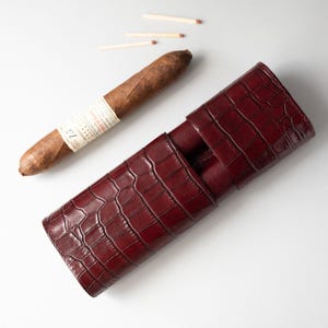 May include: A burgundy leather cigar case with a crocodile skin texture. A brown cigar with a white label rests next to the case, along with three matches. The case is designed to hold cigars and protect them.