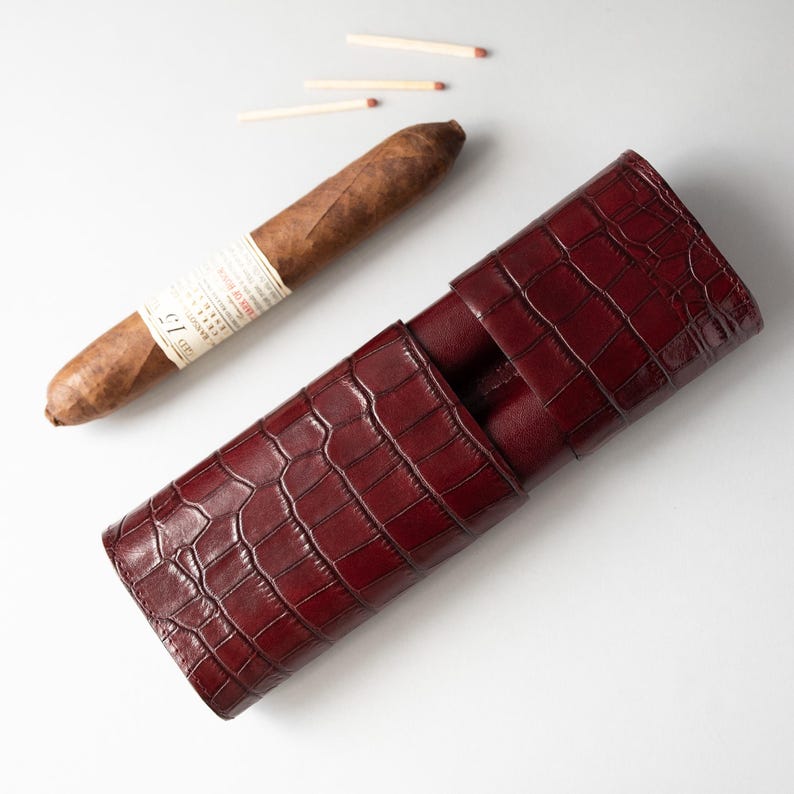 May include: A burgundy leather cigar case with a crocodile skin texture. A brown cigar with a white label rests next to the case, along with three matches. The case is designed to hold cigars and protect them.