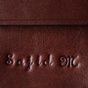 May include: A dark brown leather surface with a smooth, slightly grained texture. The name Sajid M is embossed into the material in a cursive font, creating a recessed, three-dimensional effect across the surface of the dark brown leather.
