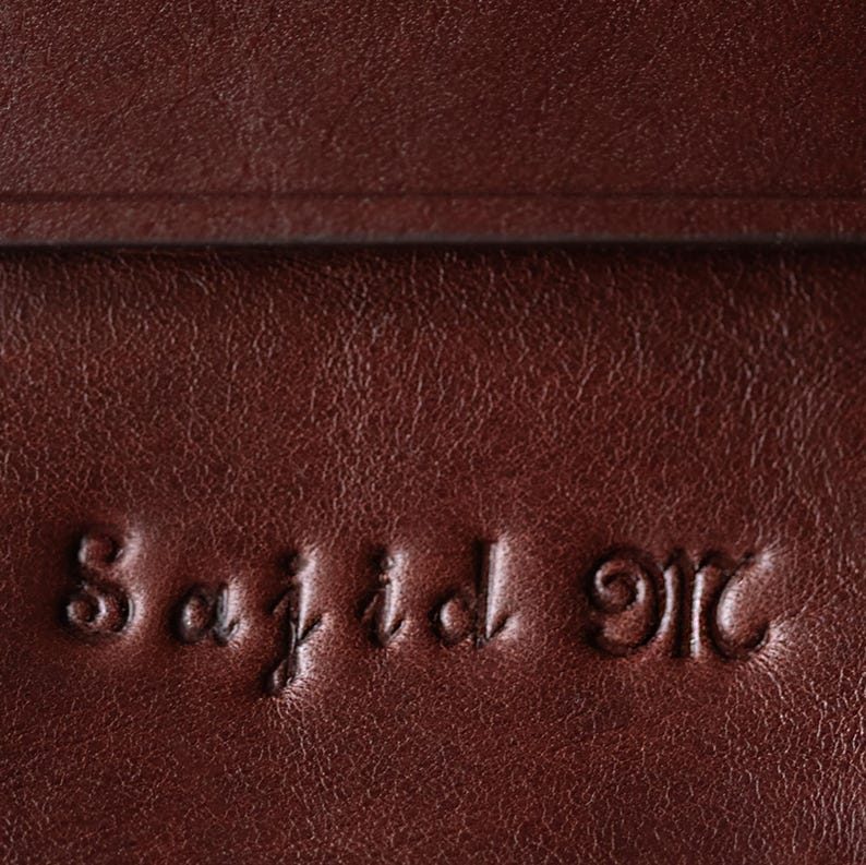 May include: A dark brown leather surface with a smooth, slightly grained texture. The name Sajid M is embossed into the material in a cursive font, creating a recessed, three-dimensional effect across the surface of the dark brown leather.