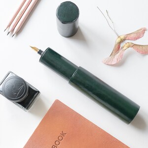 May include: A green leather pen case with a gold nib, a brown leather notebook with the word "NOTEBOOK" embossed on the cover, a bottle of black ink, and a few pencils on a white surface.