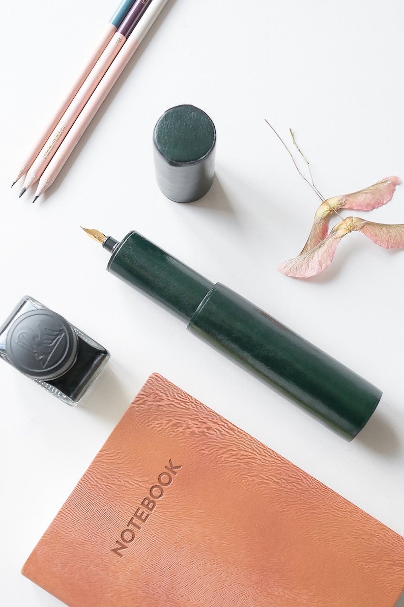 May include: A green leather pen case with a gold nib, a brown leather notebook with the word "NOTEBOOK" embossed on the cover, a bottle of black ink, and a few pencils on a white surface.