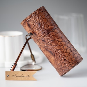 Puede incluir: Un estuche de gafas de cuero marrón con un patrón texturizado similar al de un cocodrilo. El estuche cilíndrico está en ángulo, con un par de gafas en su interior. Una pequeña etiqueta que dice "Handmade" está adjunta.