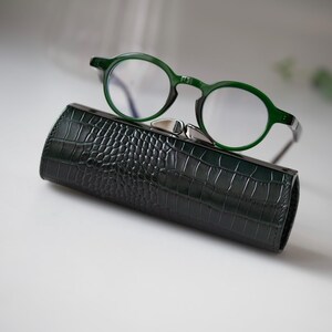 K&ouml;nnte beinhalten: Eine gr&uuml;n gerahmte Brille liegt auf einem dunkelgr&uuml;nen, strukturierten Brillenetui. Das Etui hat einen silbernen Verschluss. Die Brille hat runde Gl&auml;ser und einen dunkelgr&uuml;nen Rahmen. Das Etui scheint aus Leder zu sein.