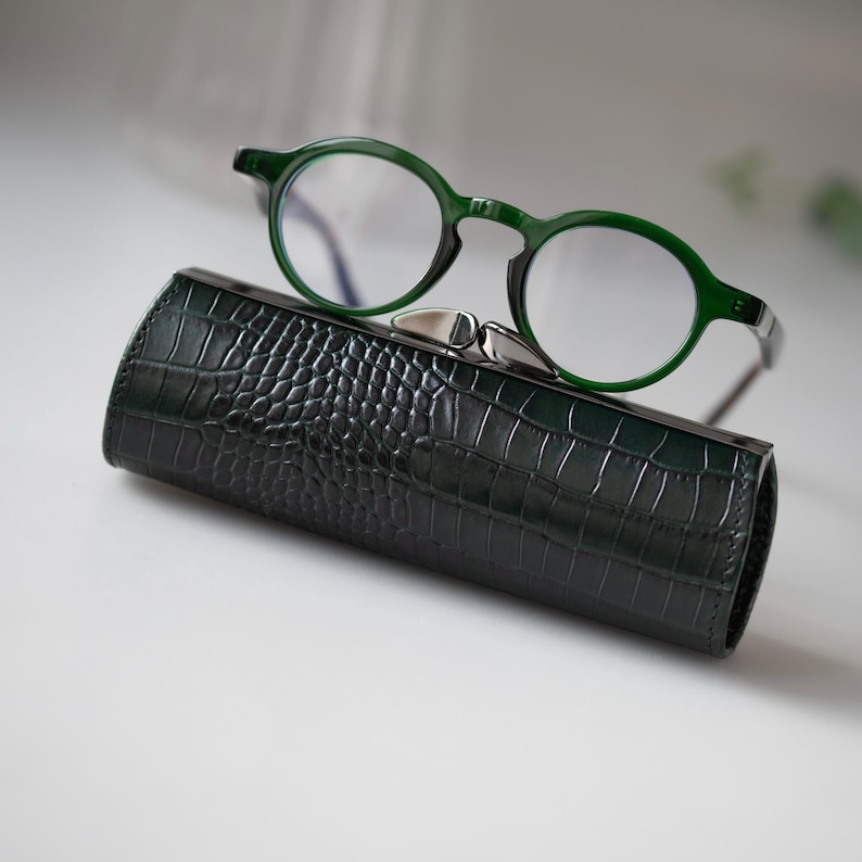 K&ouml;nnte beinhalten: Eine gr&uuml;n gerahmte Brille liegt auf einem dunkelgr&uuml;nen, strukturierten Brillenetui. Das Etui hat einen silbernen Verschluss. Die Brille hat runde Gl&auml;ser und einen dunkelgr&uuml;nen Rahmen. Das Etui scheint aus Leder zu sein.