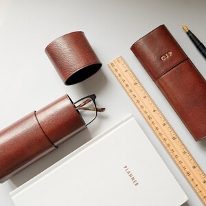 K&ouml;nnte beinhalten: Eine flache Anordnung von braunen Lederaccessoires, darunter ein Brillenetui, ein Stiftetui und zwei kleine runde Beh&auml;lter. Ein Holzlineal und ein wei&szlig;er Planer sind ebenfalls im Bild enthalten.