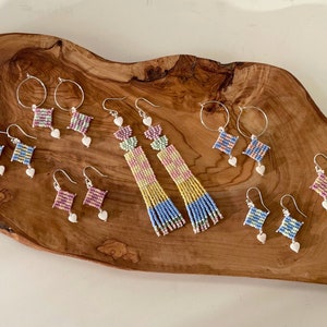 Puede incluir: Una colecci&oacute;n de pendientes de cuentas de colores con dijes de coraz&oacute;n. Los pendientes se muestran sobre una superficie de madera. Los pendientes est&aacute;n hechos con cuentas peque&ntilde;as de varios colores, incluyendo rosa, azul, amarillo y verde.