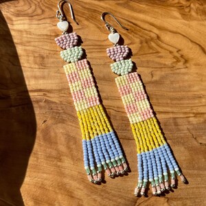 Puede incluir: Un par de pendientes largos de cuentas con un esquema de color pastel. Los pendientes presentan tres cuentas en forma de coraz&oacute;n en la parte superior, seguidas de una secci&oacute;n de cuentas rayadas y una franja de cuentas en la parte inferior. Los pendientes est&aacute;n hechos con una variedad de colores, incluyendo rosa, amarillo, azul y verde.