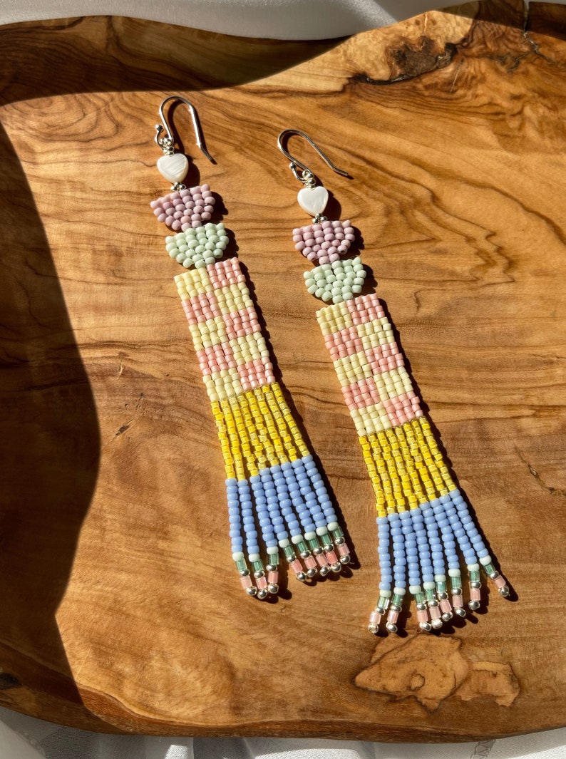 Puede incluir: Un par de pendientes largos de cuentas con un esquema de color pastel. Los pendientes presentan tres cuentas en forma de coraz&oacute;n en la parte superior, seguidas de una secci&oacute;n de cuentas rayadas y una franja de cuentas en la parte inferior. Los pendientes est&aacute;n hechos con una variedad de colores, incluyendo rosa, amarillo, azul y verde.