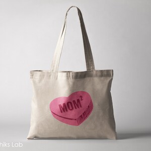 May include: A beige canvas tote bag with a pink heart-shaped design that says "MOM^2" in dark pink lettering.
