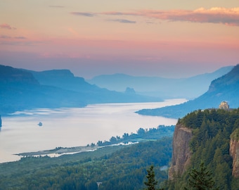 Può includere: Fotografia di un paesaggio con un ampio fiume che serpeggia attraverso una valle, fiancheggiato da montagne. Il cielo passa dal rosa e dall'arancione al blu, con un edificio in cima a una scogliera.