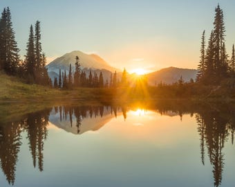 May include: A scenic landscape featuring a mountain range, a calm lake, and a vibrant sunset. The sun's rays burst through the sky, illuminating the trees and reflecting in the water. The image captures a serene natural environment.