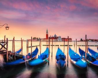 Può includere: Una vista panoramica di Venezia, Italia, all'alba con gondole attraccate a un molo. Le gondole sono blu e l'acqua è calma. Il cielo è di un rosa e un arancio vibranti.
