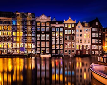 Può includere: Una vista notturna di una fila di edifici illuminati con finestre che riflettono le luci della città. Gli edifici sono allineati lungo un canale le cui acque riflettono le luci. Una piccola barca è ormeggiata nel canale.