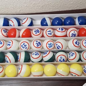 May include: A selection of colourful bingo balls displayed in a compartmentalised box. The balls are white, red, blue, green, and yellow, each with a number and letter. The numbers range from 1 to 66. The balls are for a bingo game.