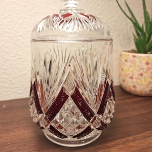 Op de afbeelding: Een heldere glazen pot met een deksel, met een diamantgeslepen patroon en een robijnrode accentkleur. De pot heeft een ronde basis en een decoratieve knop op het deksel. De pot staat op een houten oppervlak.