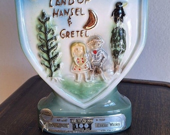May include: Vintage Beam 100 months old bourbon whiskey bottle. The ceramic bottle is shield-shaped with a light blue and green color scheme. It features a relief design of Hansel and Gretel, a crescent moon, and the word "GERMANY".
