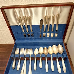 Puede incluir: Un estuche de cubiertos de madera con interior de terciopelo azul, que contiene un juego de tenedores, cuchillos, cucharas y un cuchillo de mantequilla plateados. El estuche está abierto, revelando los utensilios cuidadosamente dispuestos.