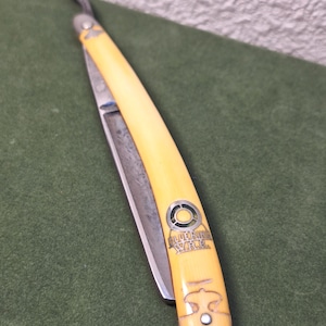 Op de afbeelding: Een vintage recht scheermes met een crèmekleurige handgreep en een zilveren lemmet. De handgreep is voorzien van de tekst "Lutering Yard" in een decoratief embleem. Het scheermes ligt op een groene achtergrond.