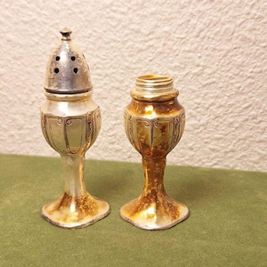 Puede incluir: Dos saleros y pimenteros antiguos plateados y dorados. Uno tiene tapa con agujeros, el otro está abierto. Ambos tienen un diseño decorativo y se asientan sobre una base de pedestal.