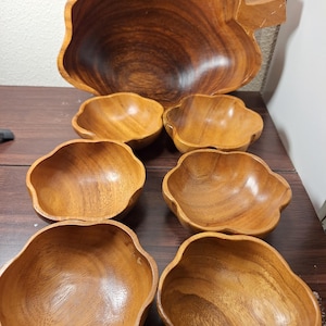 Pode incluir: Um conjunto de saladeiras de madeira com uma tigela grande em forma de folha e seis tigelas menores a condizer. As tigelas são feitas de madeira de tons quentes com padrões de grão visíveis. O conjunto está disposto numa superfície castanha escura.