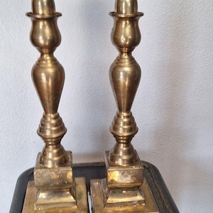 May include: Two vintage brass candlesticks with square bases and ornate, detailed stems. The candlesticks have a classic design, with a round top to hold candles. The brass has a warm, aged patina.