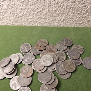 Puede incluir: Una colección de monedas antiguas de un centavo de dólar estadounidense, esparcidas sobre una superficie verde. Las monedas son de color plateado, con desgaste visible, y presentan el perfil de Abraham Lincoln. Las palabras "ONE CENT" son visibles en varias monedas.