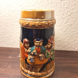 May include: A ceramic beer stein with a brown top and base, and a dark blue midsection. The stein features a relief of three figures seated around a table, holding mugs. The stein is decorated with a relief pattern.