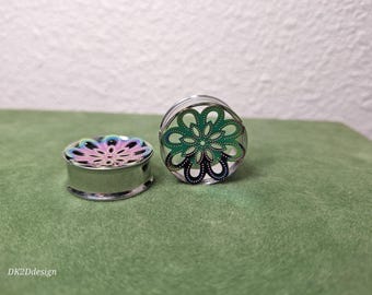 May include: Two silver-colored ear plugs with a floral design. One plug has a rainbow-colored flower, and the other has a green and black flower. The plugs are on a green surface.