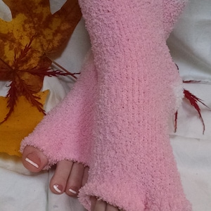 Puede incluir: Un par de calcetines rosas y peludos con ribete blanco. Los calcetines son de dedos abiertos y tienen una textura acanalada. Los calcetines se usan en los pies de una persona con esmalte de uñas blanco en los dedos de los pies.