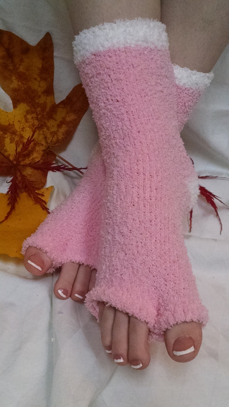 May include: A pair of pink fuzzy socks with white trim. The socks are open-toed and have a ribbed texture. The socks are worn on a person's feet with white nail polish on their toes.