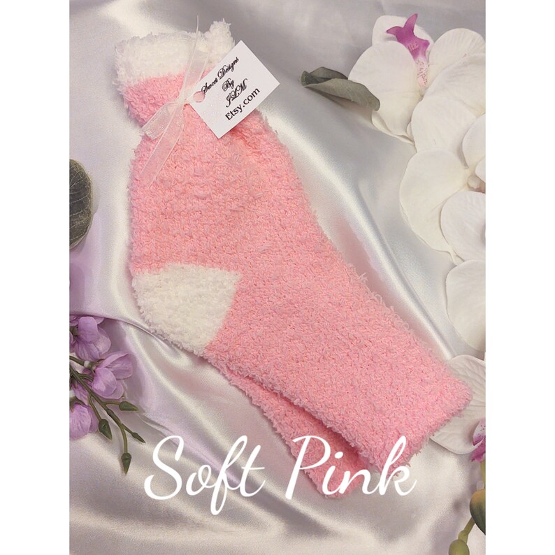 May include: A pair of soft pink fuzzy socks with a white patch on the heel. The socks are on a white satin background with a tag that says "Sweet Designs by J&M Etsy.com".