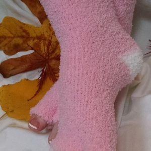May include: A pair of pink fuzzy socks with white trim. The socks are worn on a person's feet and have white nail polish on the toes.