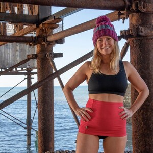 May include: A woman wearing a black sports bra and a red high-waisted skirt stands under a rusty metal structure. She is wearing a pink and grey knit hat with a pom-pom on top. The woman is smiling and looking at the camera.