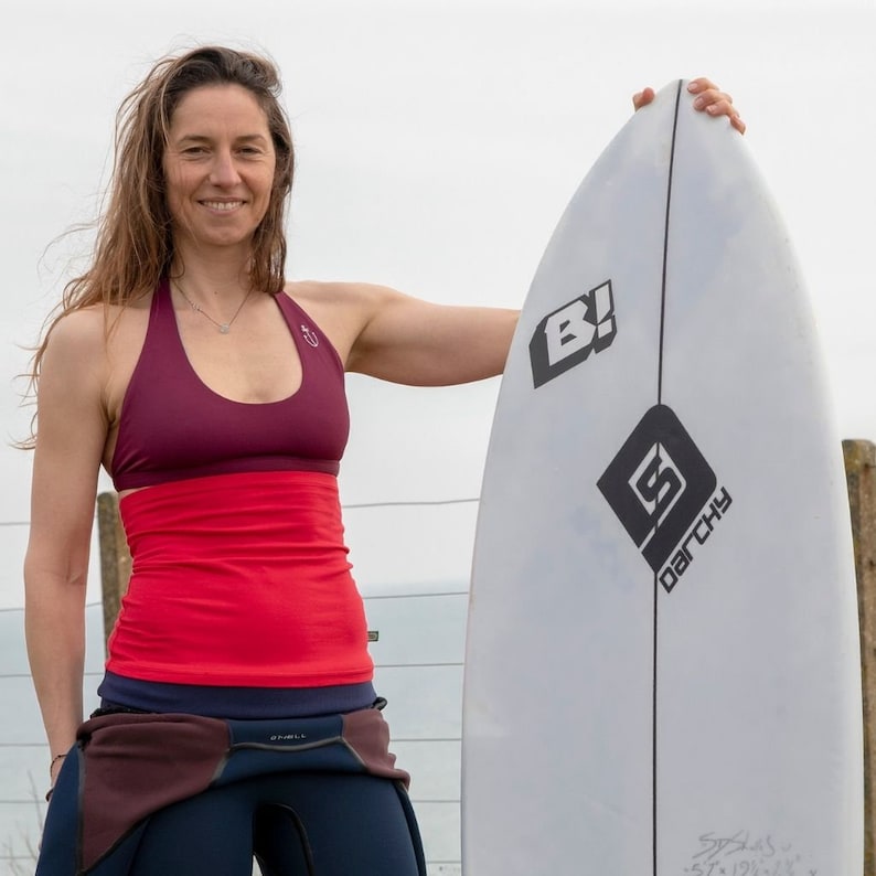 May include: A woman wearing a red and burgundy halter top and a black wetsuit stands holding a white surfboard with black lettering that says "B!" and "DACHSU" in a diamond shape.