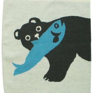 May include: A light blue and cream-colored knitted towel featuring a black bear with a blue fish in its mouth. The bear has white eyes and a cream-colored border.