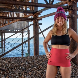May include: A woman wearing a pink skirt, black sports bra, and a pink and grey beanie stands under a rusty metal pier. The woman is smiling and looking at the camera.