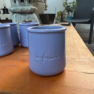 以下が含まれることがあります： 側面に「la fermiere」の文字がエンボス加工された、薄紫色のセラミックジャーがいくつかあります。ジャーは円筒形で、縁がわずかに広がっています。木製のテーブルの上に置かれています。
