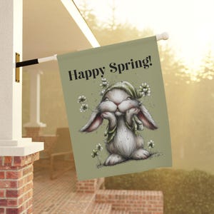 May include: A decorative flag featuring a cute bunny illustration with the text "Happy Spring!". The bunny is white with long ears and is wearing a green bonnet and surrounded by flowers. The flag is a light green color.