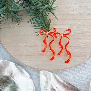 May include: Two red enamel bow-shaped ornaments with gold trim, displayed on a light wood surface with a sprig of green pine. The bows have a glossy finish and are likely Christmas decorations.