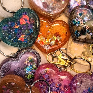 May include: A collection of 10 heart-shaped keychains with different colors and patterns. Each keychain features a clear resin heart filled with glitter, sequins, and other decorative elements. The keychains are attached to silver metal rings.
