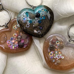 May include: Three heart-shaped keychains with different colored glitter and seashells inside. The keychains are made of clear resin and have silver key rings.