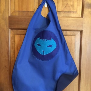 May include: A blue cape with a dark blue lining and a circular emblem featuring a teal mask design. The cape is hanging on a white hook against a wooden door.