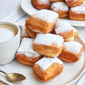 Könnte beinhalten: Ein Teller mit goldbraunen, mit Puderzucker bestäubten Beignets. Eine Tasse Kaffee mit cremiger Oberfläche und ein kleiner goldener Löffel liegen neben dem Teller. Weitere Beignets befinden sich auf einem zweiten Teller im Hintergrund.